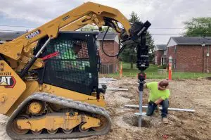 Construction worker guiding a helical pier installation using a yellow Caterpillar skid-steer loader with an auger attachment on a residential construction site.