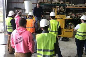 A group of construction workers or students in safety gear attending a presentation or training session in an industrial setting.