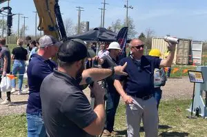 A man in a blue shirt holds up a small cylindrical part for a crowd of people, while another man films the demonstration at an outdoor training event with construction equipment in the background.