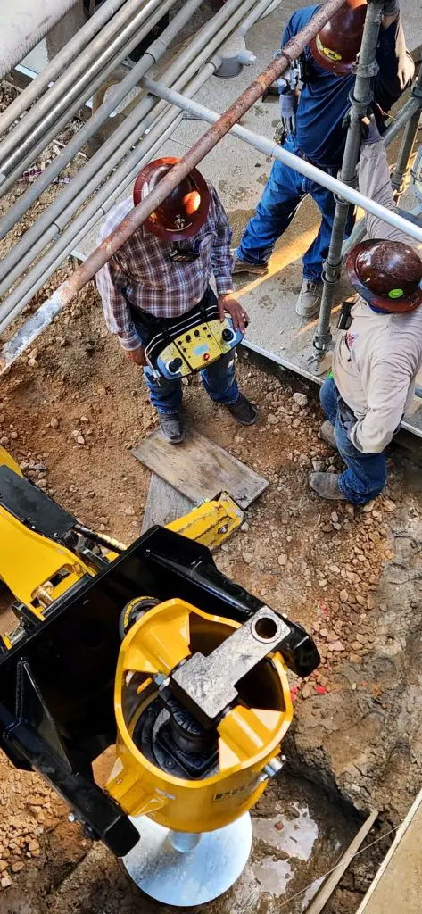 Three construction workers operating a yellow drilling or anchoring machine with a remote control on a job site with scaffolding.