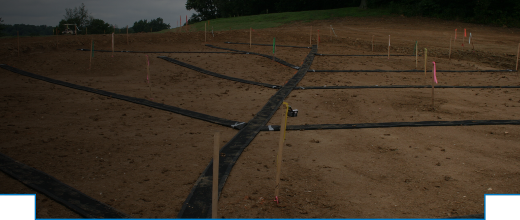 Construction site with bare earth, black silt fencing, and survey stakes marking out the foundation or utility lines.