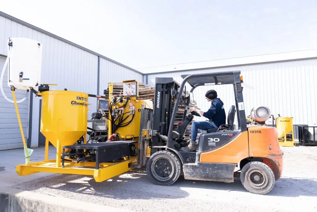 A person operating an orange Doosan forklift next to a large, yellow IN-TECH ChemGrout industrial mixing machine outside a white warehouse building.