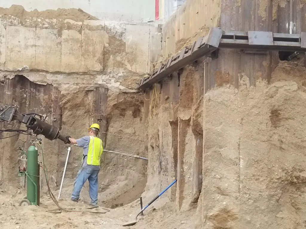 Construction worker in a yellow vest operating a large drilling or anchoring machine in a deep excavation pit with shored walls.