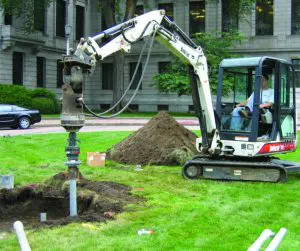 A small white excavator or mini-digger is installing a helical pile or anchor into the ground on a grassy area, with a pile of dirt next to the machine and a large building in the background.