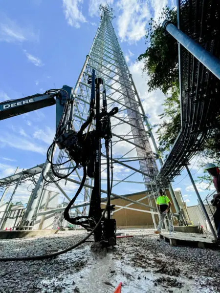 A construction worker and heavy machinery near the base of a large lattice tower, illustrating the installation of Magnacore micropiles for the tower's foundation.