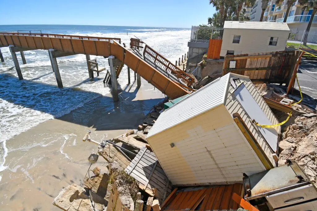 A small white shed and wooden fence have collapsed onto a sandy beach after a seawall failed, with a wooden pier and stairs partially damaged in the background.