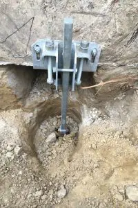 Close-up of a metal foundation underpinning bracket bolted to a concrete wall with a vertical pier extending into the excavated ground.