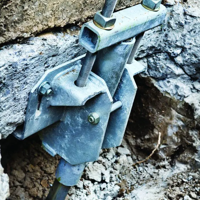 Close-up of a foundation underpinning bracket, showing a galvanized steel plate bolted to a rock foundation with threaded rods and a steel pipe extending into the soil.