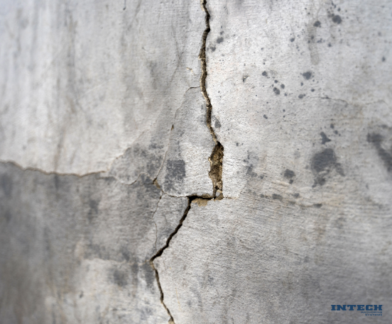 Close-up of a deep, vertical crack in a weathered gray concrete or stucco wall, with a horizontal crack intersecting it.