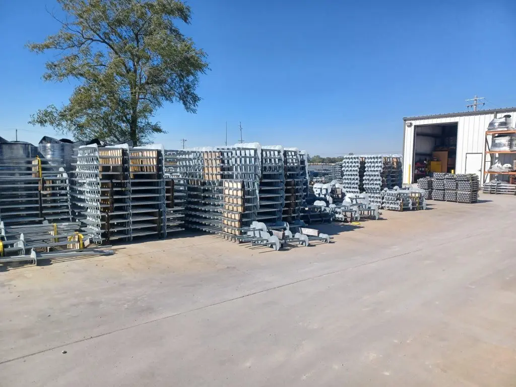Outdoor storage yard with large stacks of metal helical piles and anchoring components on pallets, next to a white warehouse building under a clear blue sky.