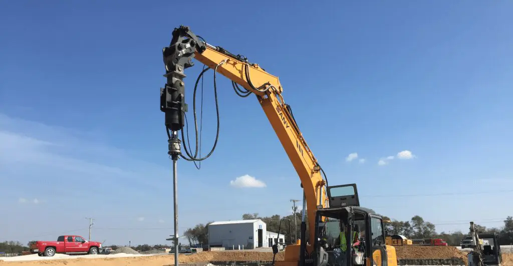 Yellow SANY excavator with a drilling rig attachment installing a ground anchor at a construction site.