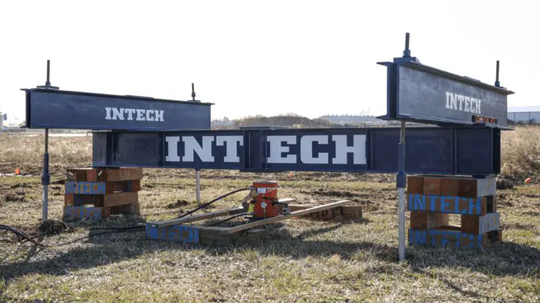 Industrial scene showing a large clamshell grab bucket suspended by cables, descending into a dark, concrete structure, possibly for loading or unloading bulk material like coal, with steam or dust visible on the right.