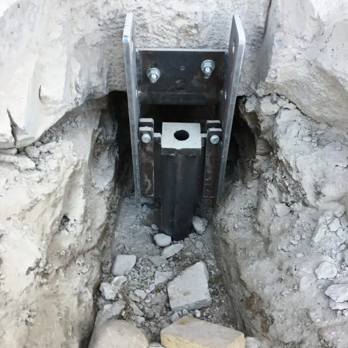 A close-up view of a steel foundation anchor, possibly a helical or resistance pier, installed in a trench of dirt and rocks, showing the bracket and bolts connecting it to the foundation.