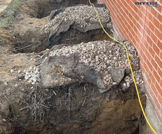 Excavated trench next to a brick foundation, showing two layers of gravel and geotextile fabric for drainage or underpinning.