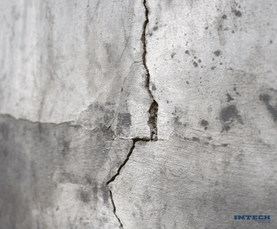 Close-up of a deep, vertical crack in a weathered gray concrete or stucco wall, with a horizontal crack intersecting it.