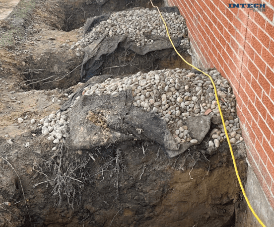 Excavated trench next to a brick foundation, showing two layers of gravel and geotextile fabric for drainage or underpinning.